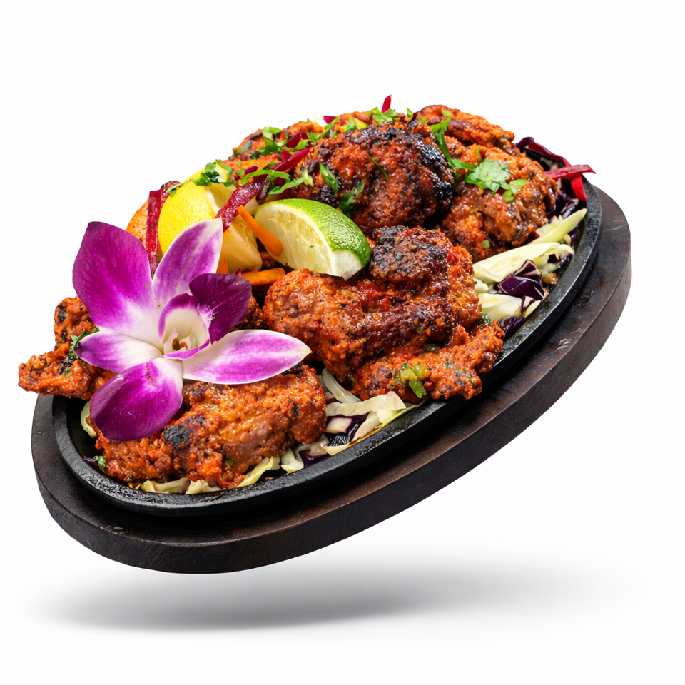 Grilled spicy meat pieces with cabbage, citrus wedges, herbs, and orchid garnish on a black sizzling platter.