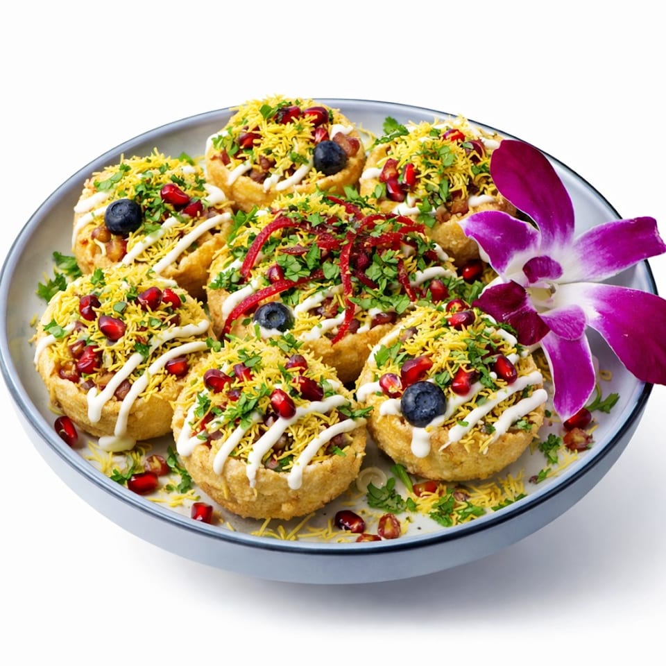 A plate of colorful Indian chaat, garnished with pomegranate, blueberries, herbs, and an orchid.