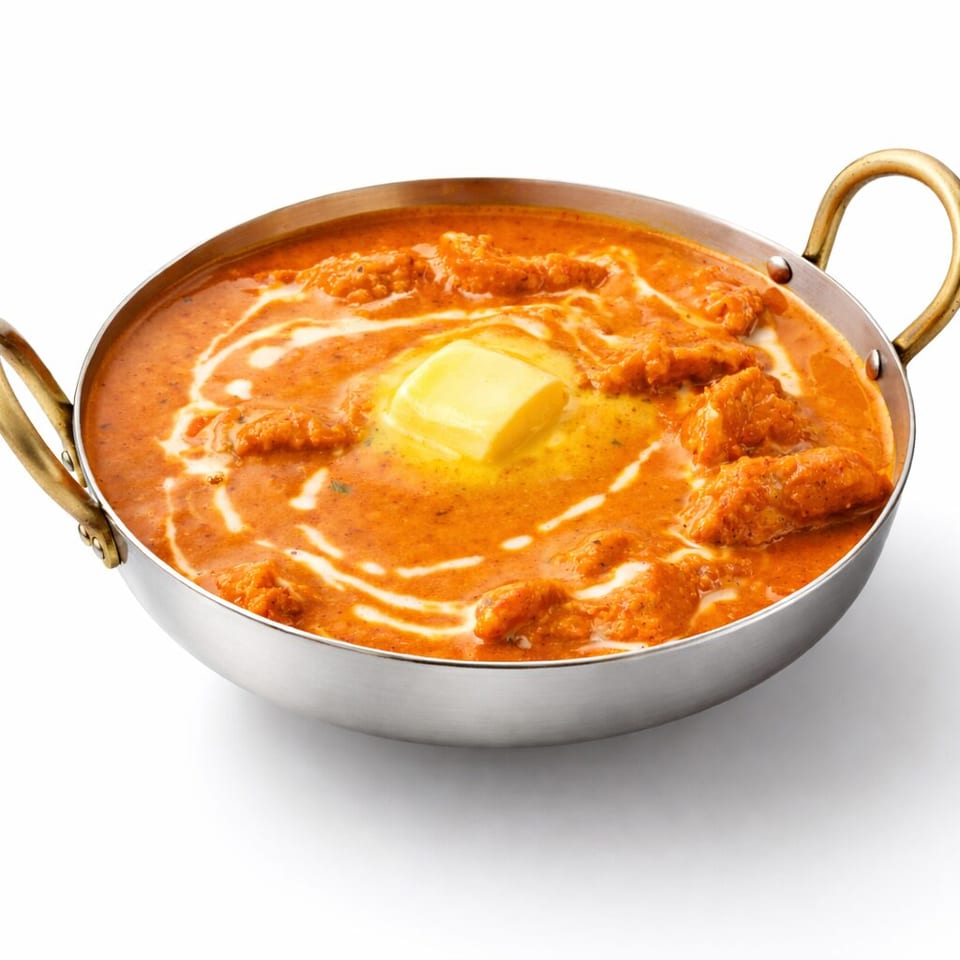 Indian butter chicken in creamy orange curry sauce, topped with butter and cream, served in a handled metal pan.
