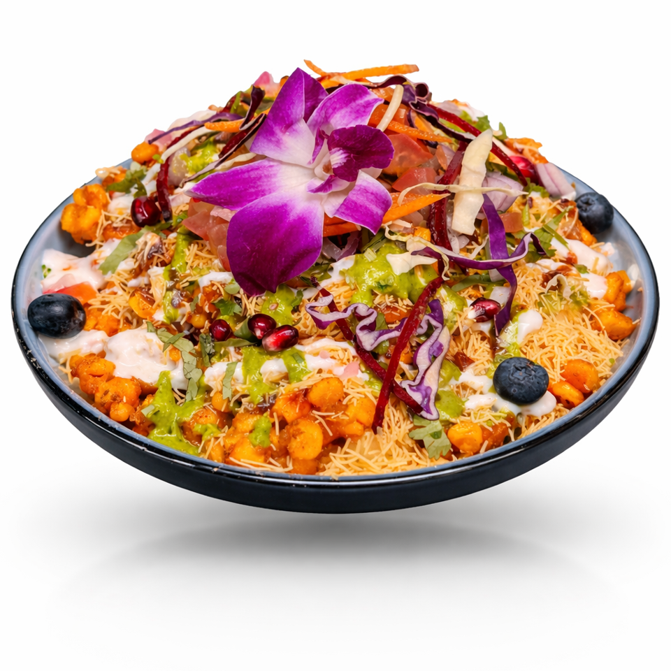 Colorful Indian chaat platter topped with sev, chutneys, vegetables, blueberries, and a purple edible flower in a black bowl.